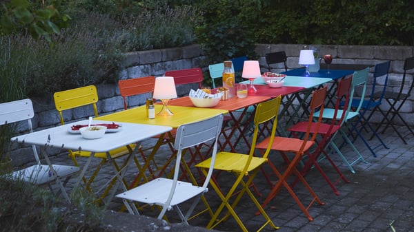 Several SUNDSÖ folding tables and chairs in different colours placed to a long outdoor table.