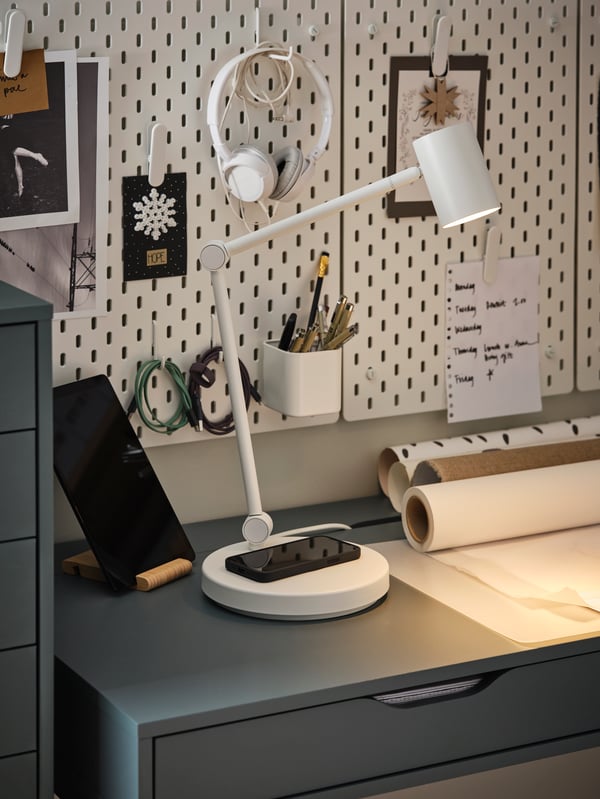 A white work lamp with wireless charging, a black phone, white pegboards, a pair of headphones, a grey-turquoise desk.