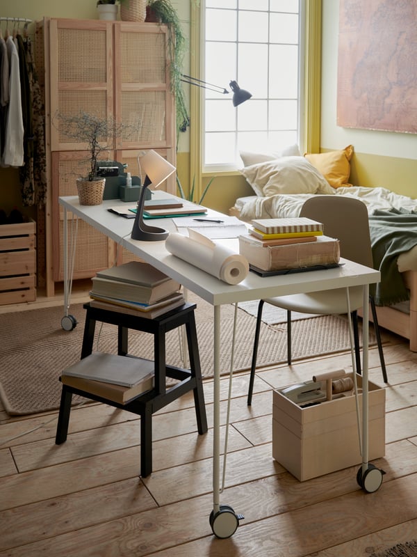 A white desk on wheels is placed in focus in a relaxed student room with a wooden storage box on three levels on the floor.