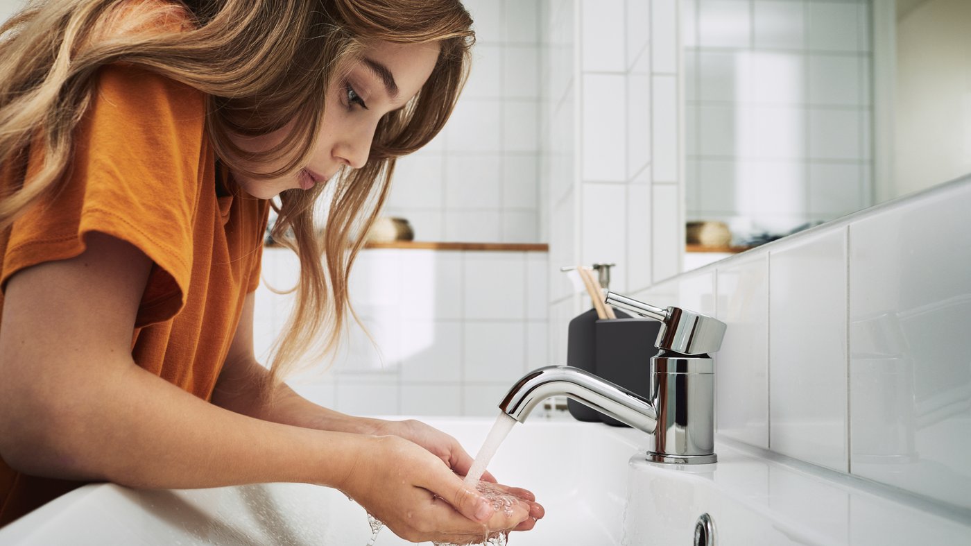 Girl drinks water out of her hands from the faucet wearing an orange t-shirt.