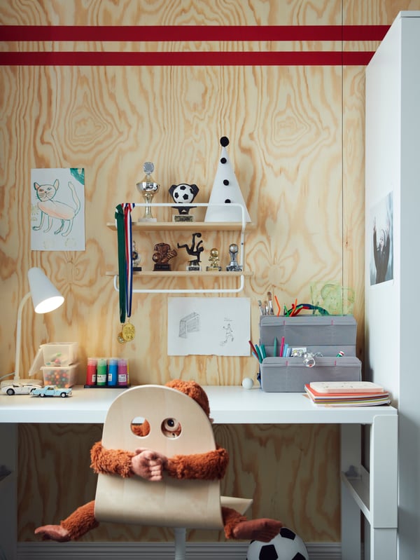 A SPORTSLIG wall shelf on a plywood wall holding trophies, medals and objects, above a desk with pens and paints on it.