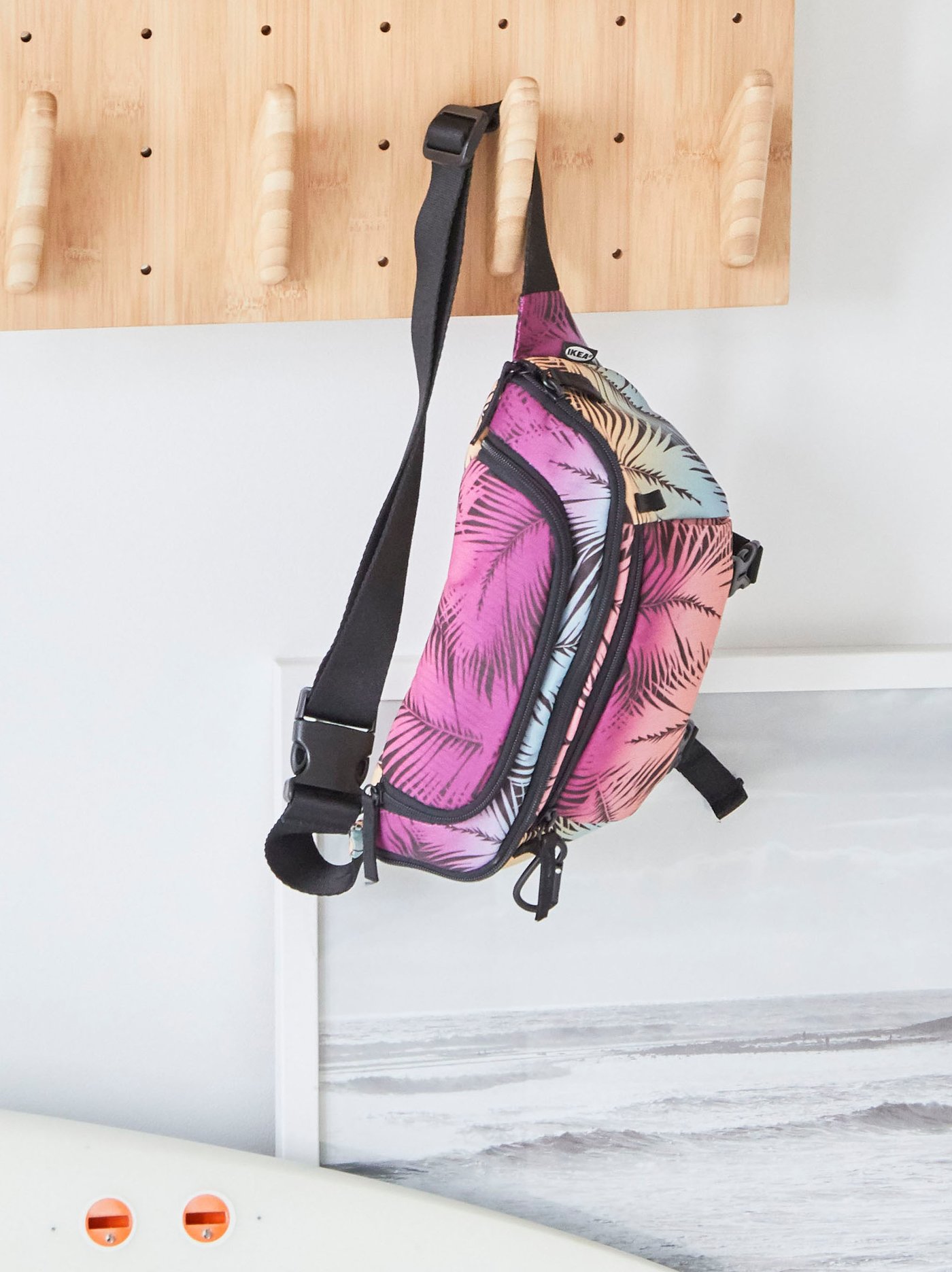 KÅSEBERGA belt bag in a colourful palm-inspired print hanging on the hook of a bamboo pegboard against a white wall.