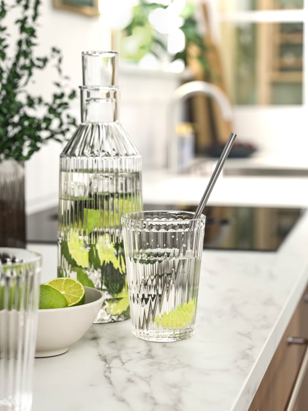 A bottle and a glass filled with water and lime wedges on an EKBACKEN worktop with a white lively marble effect/laminate.