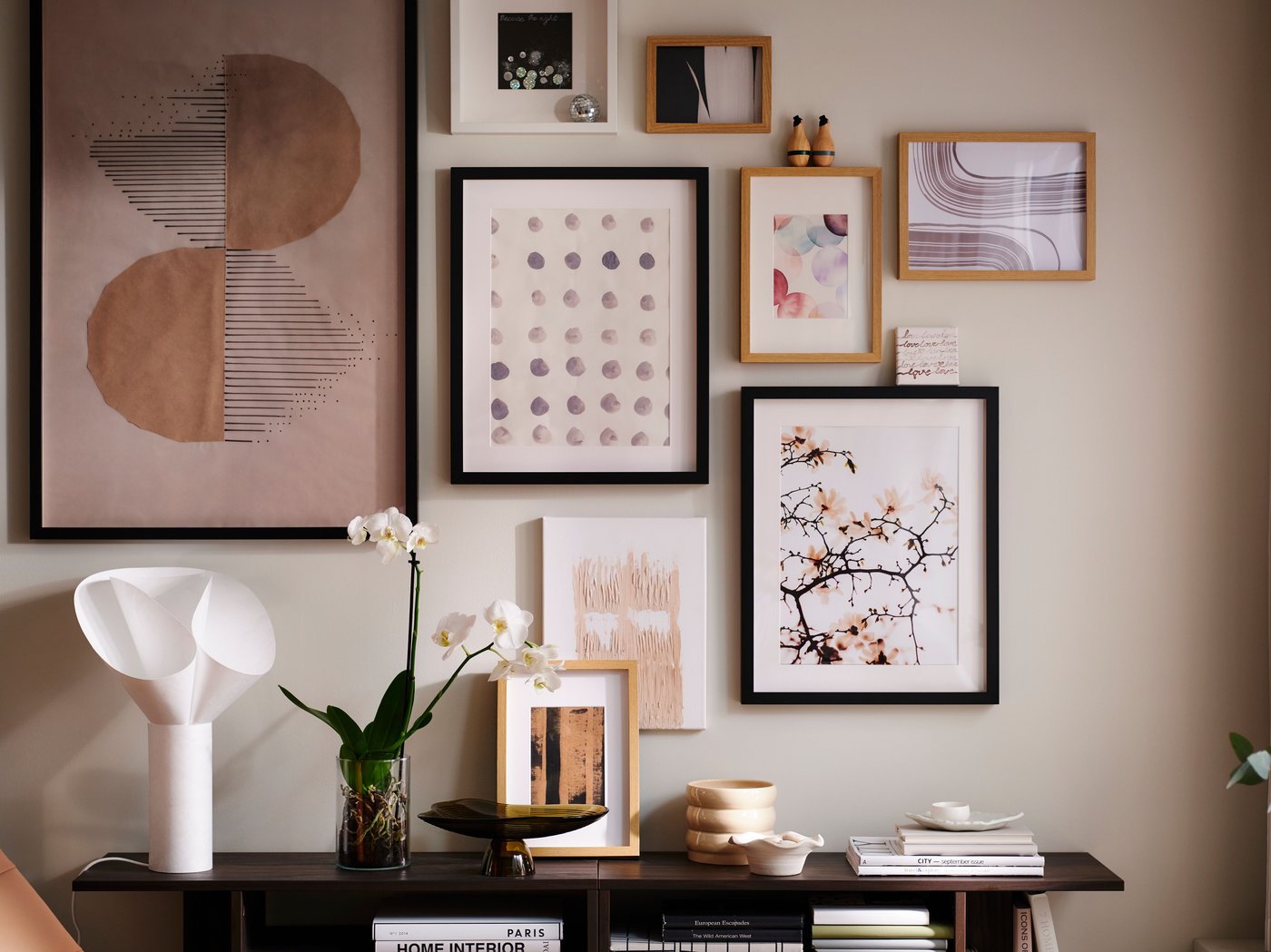A wall in a light living room with an art cluster in various RÖDALM frames above HOLMERUD side tables with orchids and deco.