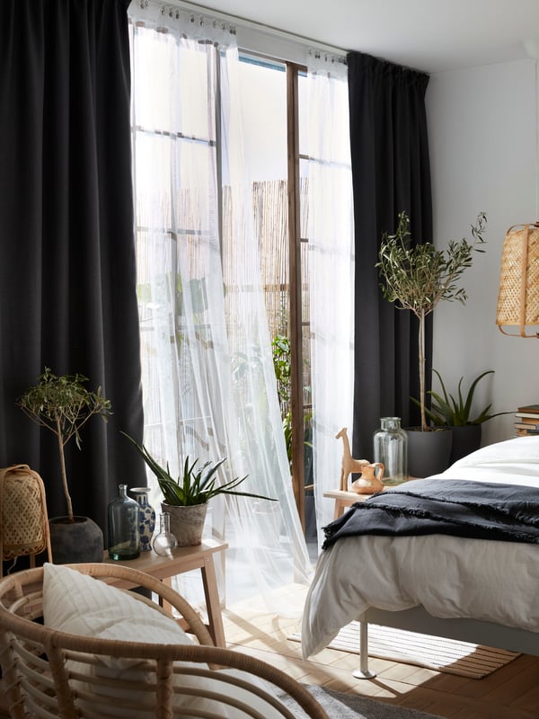 Un rideau filet blanc LILL flotte au vent devant une fenêtre ouverte dans une chambre à coucher pendant une journée ensoleillée. Il touche presque le lit.