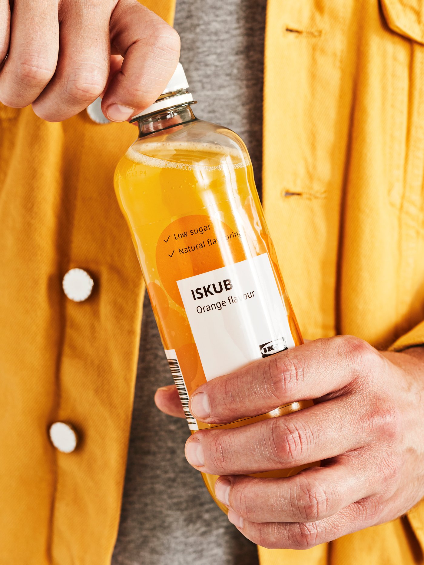 A person wearing a yellow jacket and grey jumper holding a bottle of ISKUB carbonated soft drink in orange flavour.