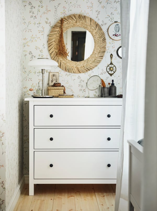A white HEMNES chest of drawers with a white table lamp on it, in front of a floral wall with a VRIGSTAD mirror on it.