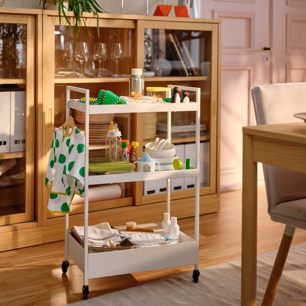 Dans une salle à manger, une desserte NISSAFORS blanche est chargée de tout le matériel nécessaire pour nourrir un bébé : un bavoir, des assiettes, des bols et des cuillères BÖRJA, etc.