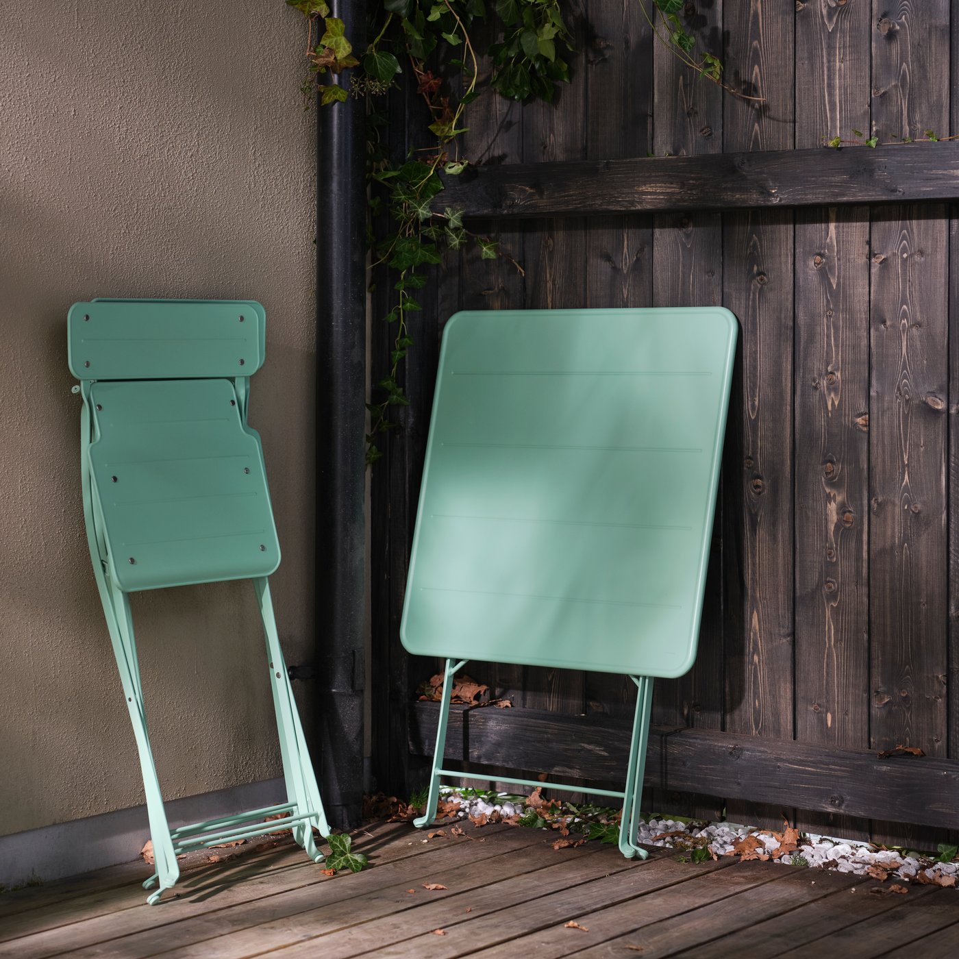 Una mesa SUNDSÖ plegable y una silla plegable SUNDSÖ plegable apoyadas contra una valla y una pared en una terraza.
