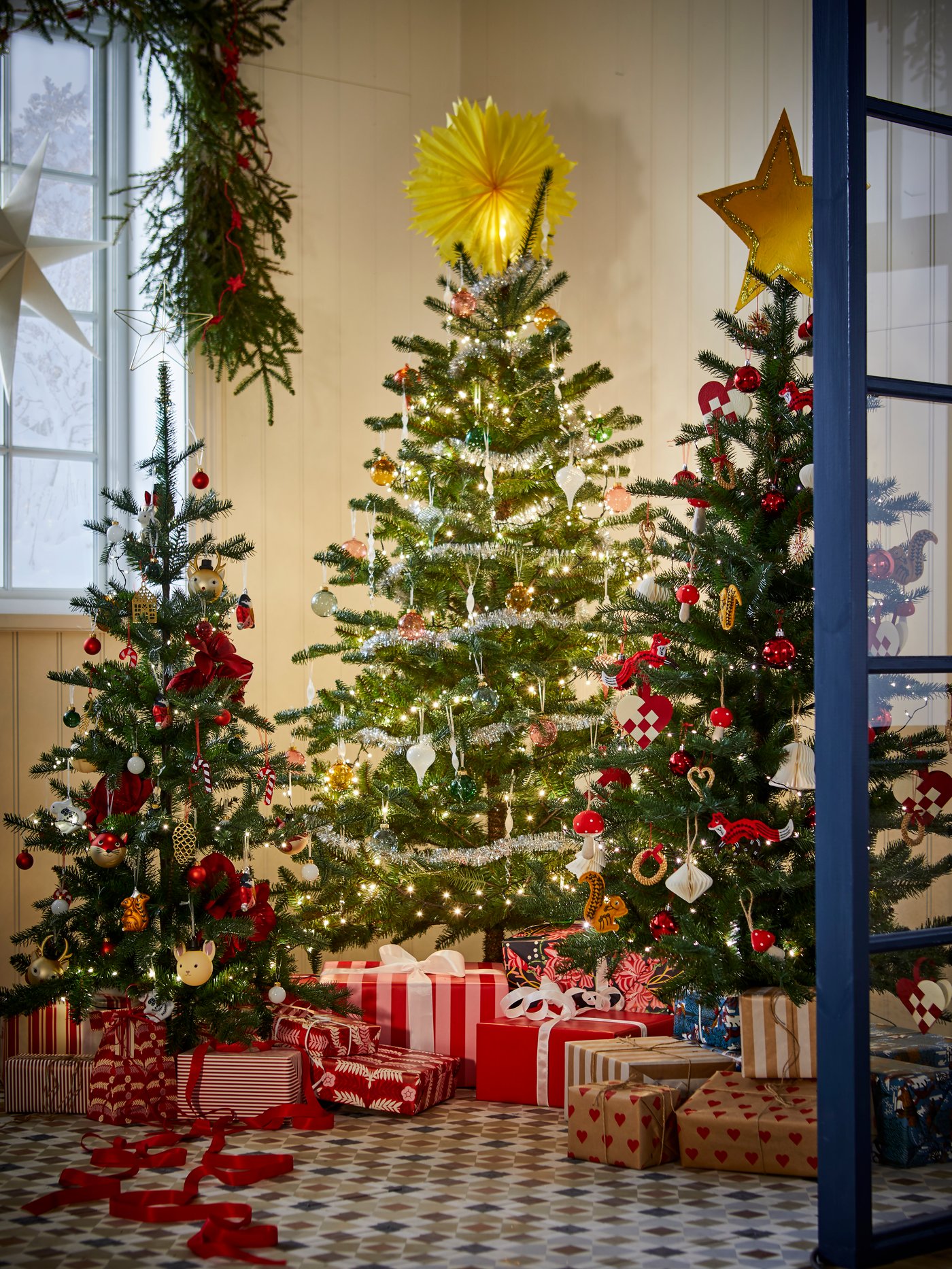 VINTERFINT artificial Christmas trees in three sizes decorated with tree ornaments, with gift wrapped presents underneath.