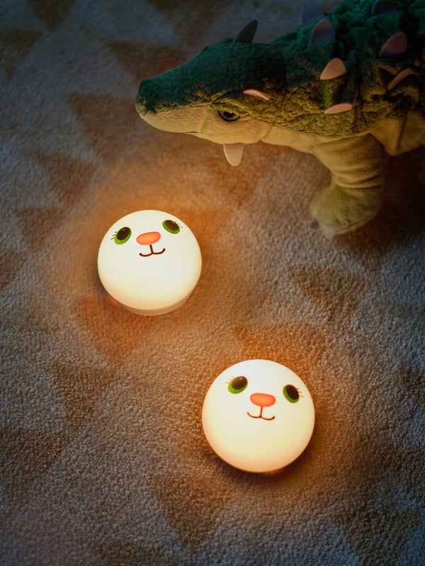 A brown rug, a JÄTTELIK dinosaur soft toy and two white KORNSNÖ LED night lights with a cute rabbit face.