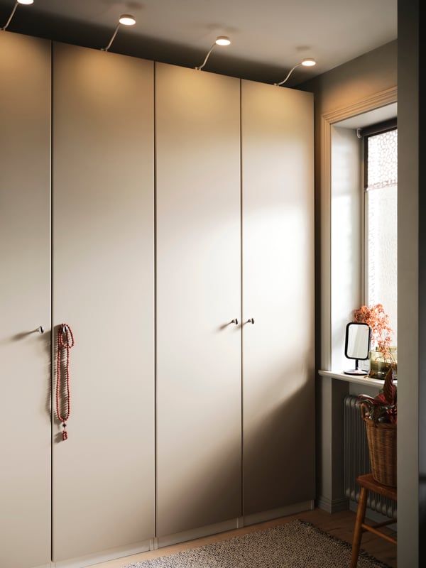 Ein beiger PAX/REINSVOLL Kleiderschrank neben einem Fenster. Entlang der Oberkante verläuft eine YTBERG Schrankbeleuchtung.