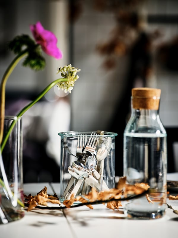 A tabletop with an IKEA 365+ clear-glass carafe with a cork, cutlery gathered in a glass jar, and two flowers in a vase.