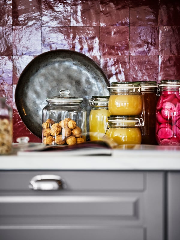 Pickles and preserves in various KORKEN and VARDAGEN jars, on a white marble effect worktop in a kitchen with red wall tiles.