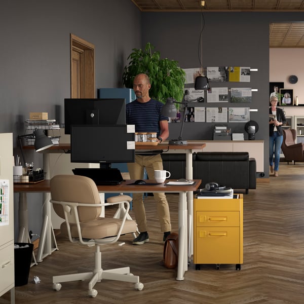 En una oficina hay unhombre trabajando de pie frente a un escritorio de altura regulable IKEA.