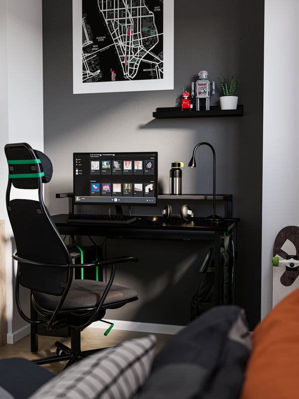 Corner of a room with a dark grey wall, a black UTVISNING desk with computer on top and a black LÖPARBANA gaming chair.