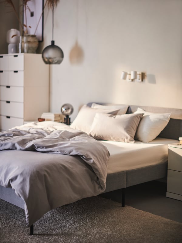 A light grey SLATTUM upholstered bed frame with an ÅKREHAMN foam mattress and NATTSVÄRMARE bed linen stands against a wall.