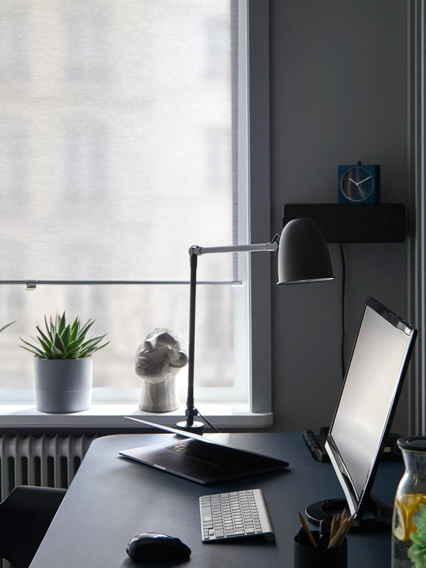 Una finestra con tende a rullo SKOGSKLÖVER bianche quasi completamente abbassate accanto a una scrivania con una lampada e lo schermo di un computer - IKEA