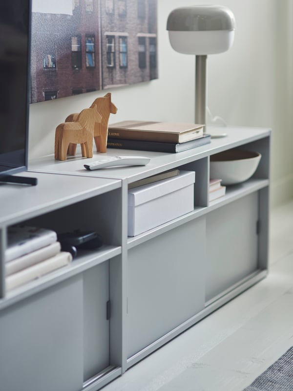 Two light grey SPIKSMED TV benches have a TV unit, remote control, wooden horse figures, books and a table lamp on top.