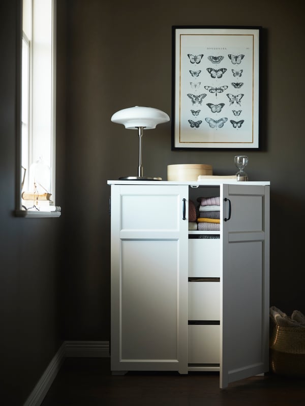 A white GREÅKER cabinet with drawers with one door open and folded clothes on the shelf inside, beneath a butterfly poster.