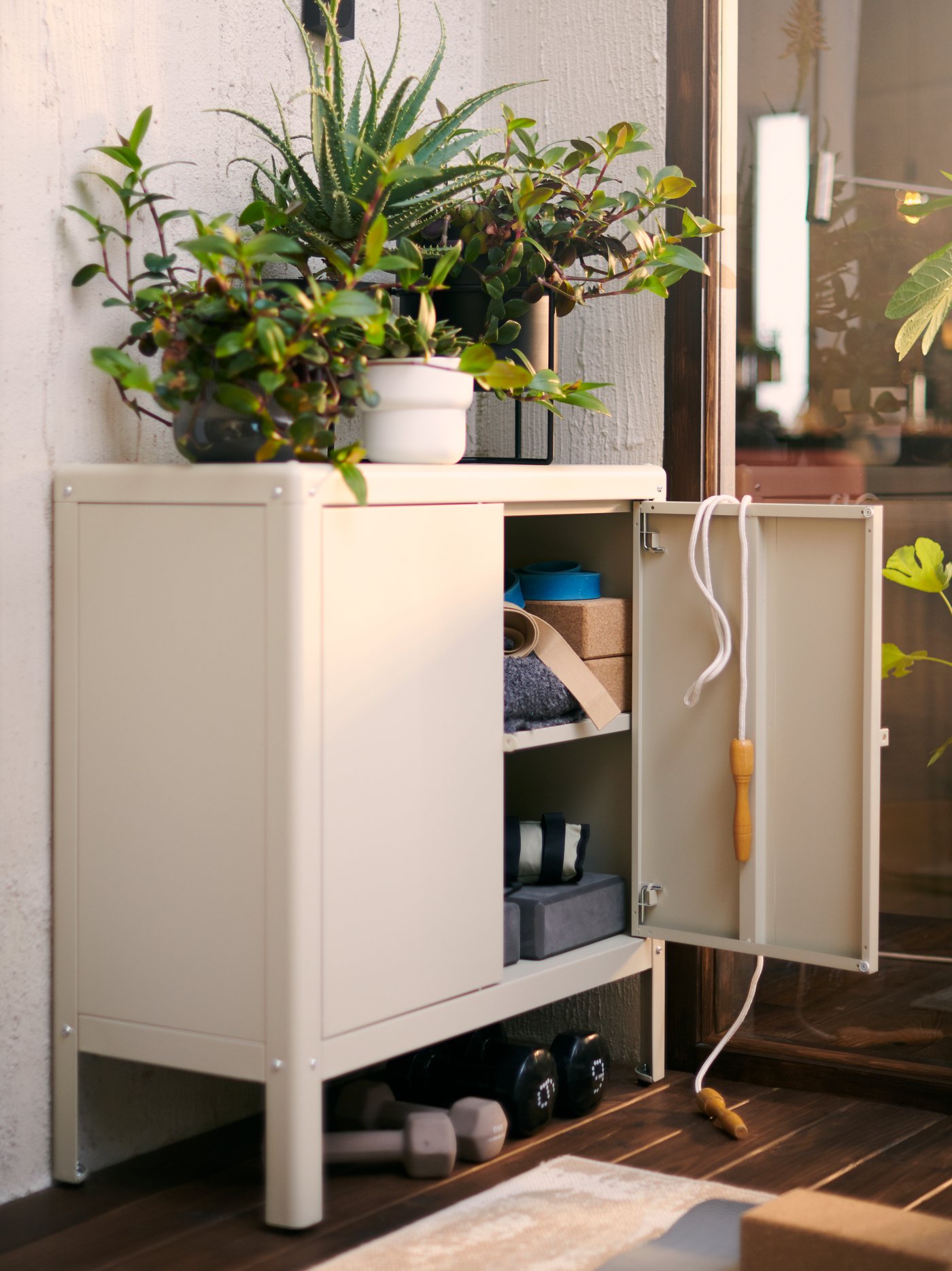 A beige KOLBJÖRN cabinet with one door open and boxes on shelves inside. Plants are on top and hand weights are underneath.