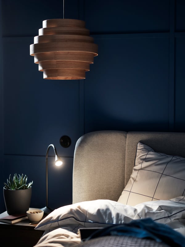 The top of a TUFJORD upholstered bed with a NÄVLINGE LED work lamp and a plant in a PERSILLADE pot on a bedside table by it.