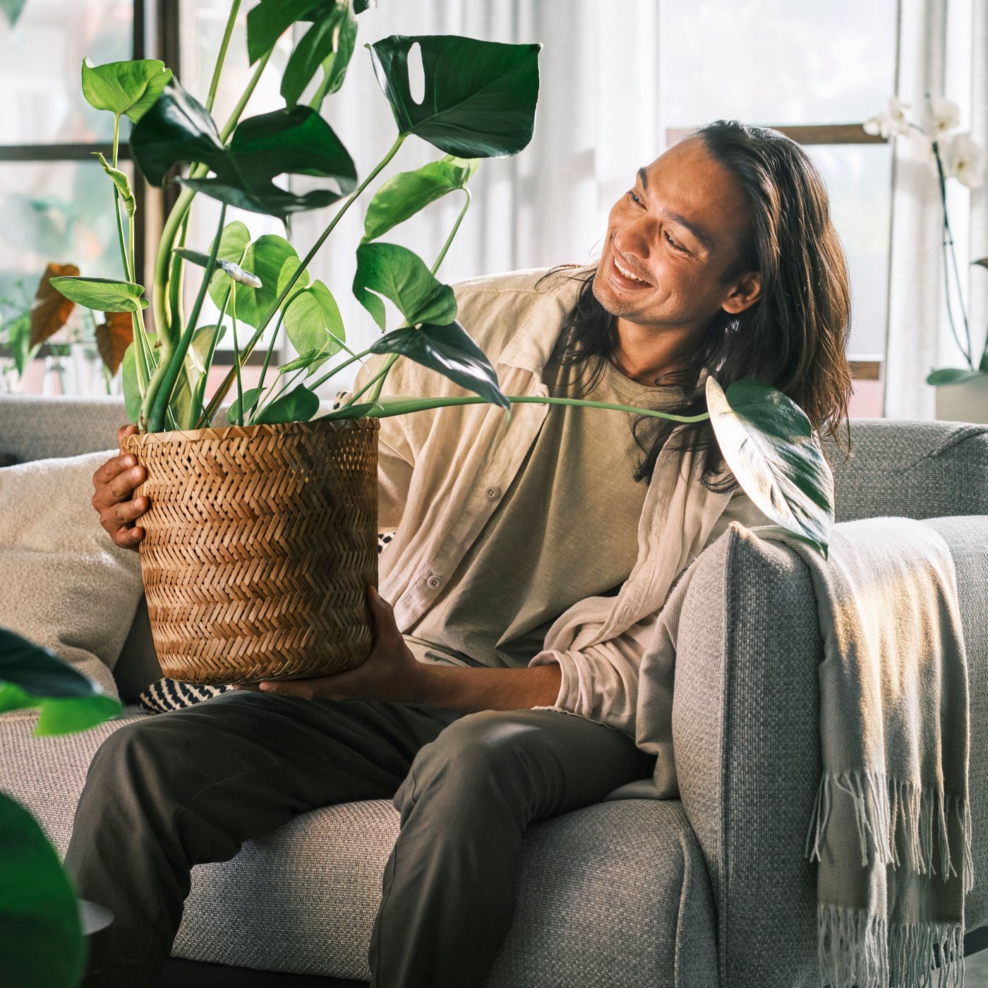 Una persona sorridente seduta su un divano a 3 posti ÄPPLARYD grigio chiaro; in mano ha una monstera piantata in un portavasi in bambù KLYNNON.