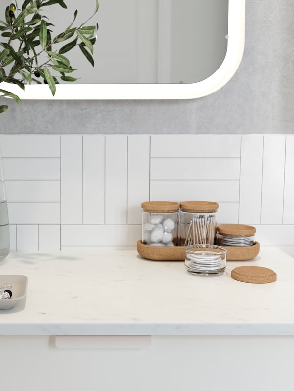 Cotton swabs and cotton wool balls in glass jars with cork lids stand on a wash-basin with white marble effect.
