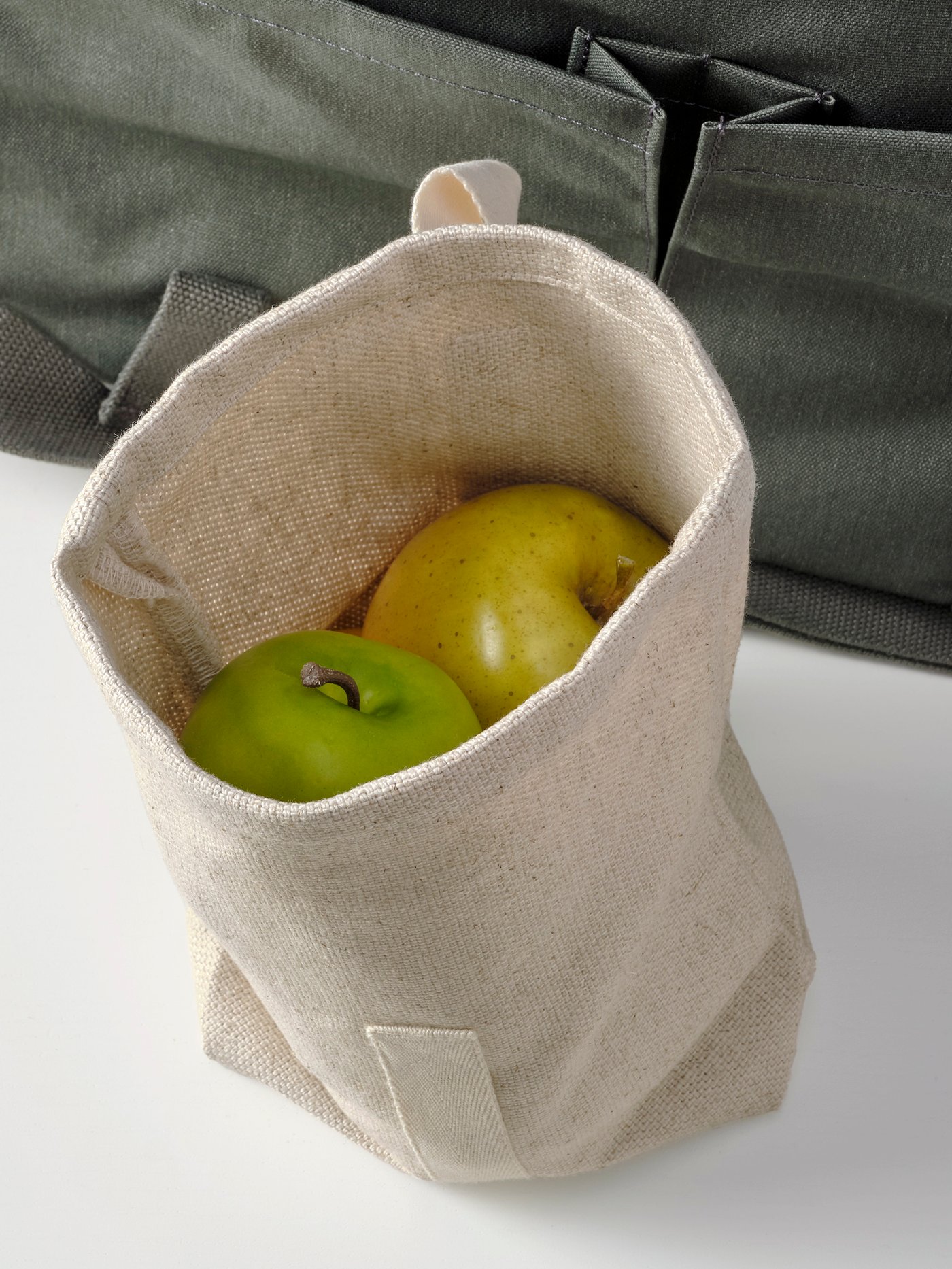 An open HAJMAL snack bag in natural fabric containing two fresh green apples in front of a bag on a white surface.