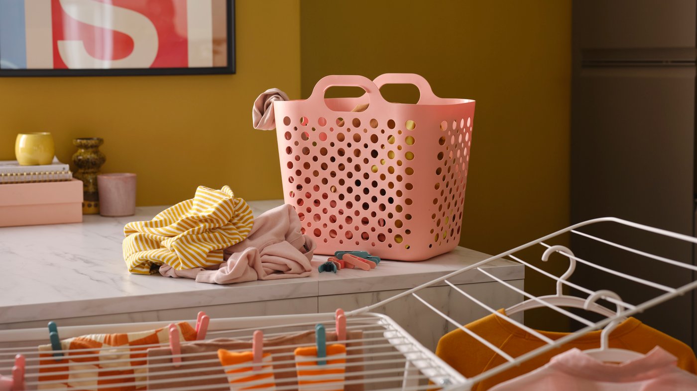 A pink SLIBB flexible laundry basket and a pile of clothes on a kitchen worktop, plus a white drying rack with more clothes.
