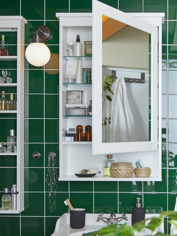 A green tiled wall with a wall-mounted HEMNES mirror cabinet that is open, revealing beauty products and accessories.