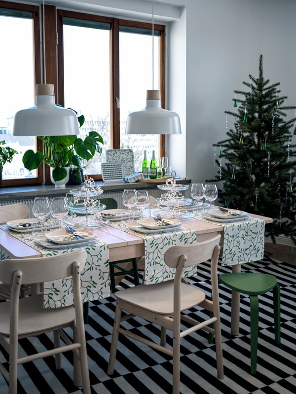 Una mesa extensible RÖNNINGE de abedul con cristalería y vajilla, con sillas RÖNNINGE a juego junto a un árbol de navidad.