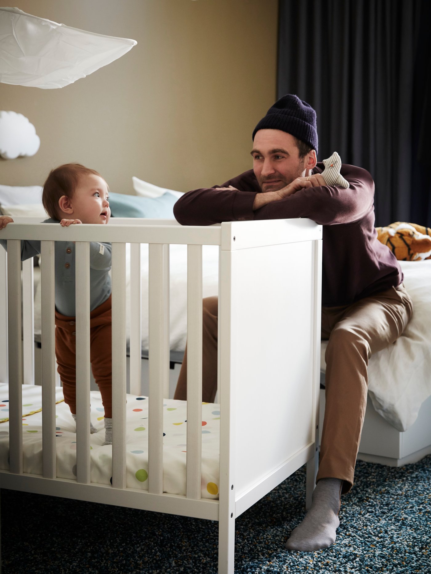 A man holding a soft toy looking at a child who is standing up in a SUNDVIK white cot.