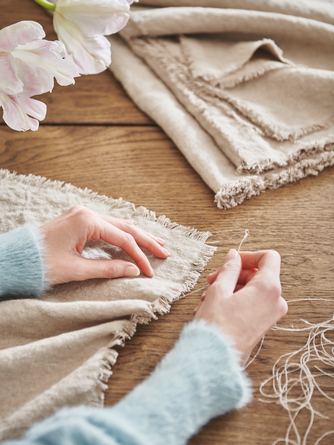 A pair of hands decoratively fraying a length of natural-colour AINA fabric by pulling threads from its edges.