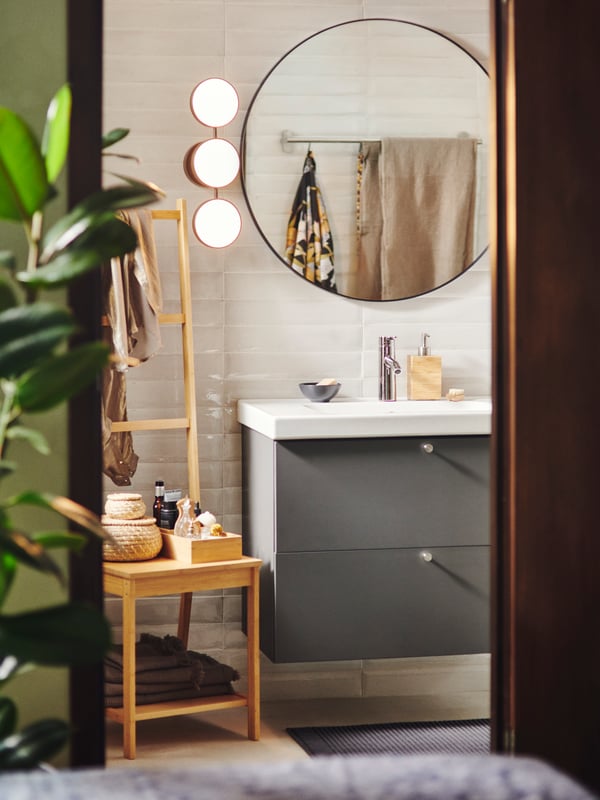 A GODMORGON washstand is mounted on a wall below a round LINDBYN mirror. A RÅGRUND storage chair is next to it.