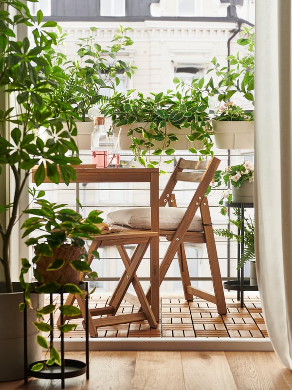 A light-brown-stained NÄMMARÖ outdoor table, foldable chair and foldable stool on a balcony enveloped in leafy potted plants.
