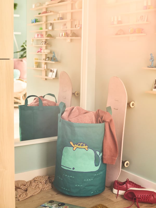 A BLÅVINGAD storage bag with a drawing of a whale and a turtle is in a corner alongside a skateboard in front of a mirror.