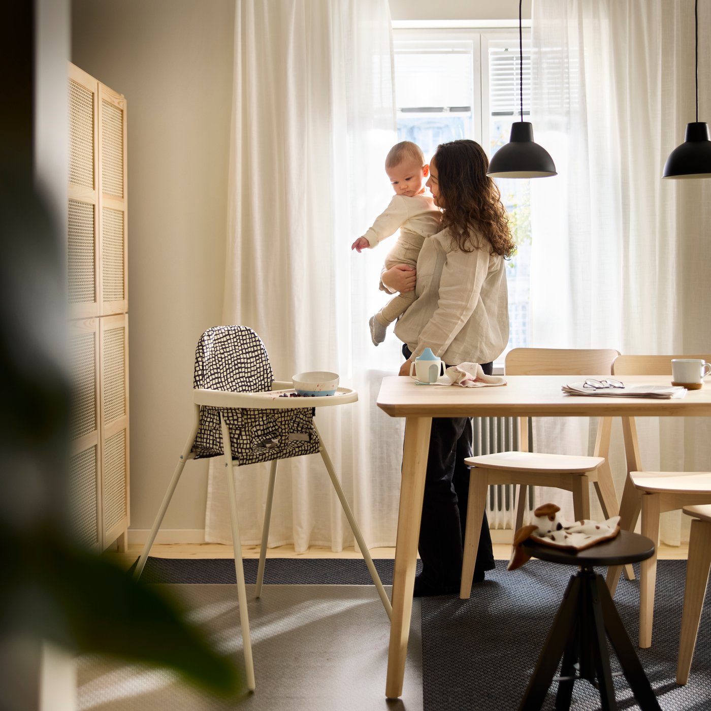 SANDKATT highchair placed beside a dining table in a bright room where an adult is holding a baby.