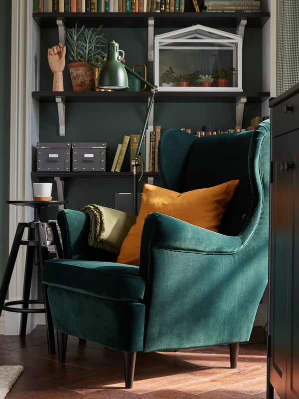 A green STRANDMON wing chair and a black DALFRED bar stool stand in front of shelves which hold books, boxes and plants.