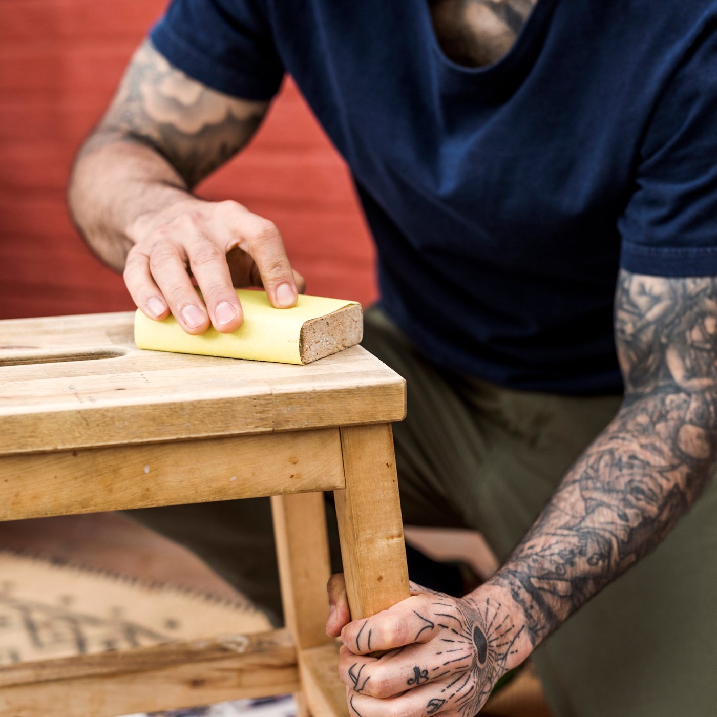 En person med tatueringar på armarna använder sandpapper för att slipa en pall i trä.