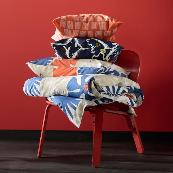 An easy chair sitting by a red wall with a duvet and pillow in HYLSNEJLIKA bed linen stacked on it, and two cushions on top.