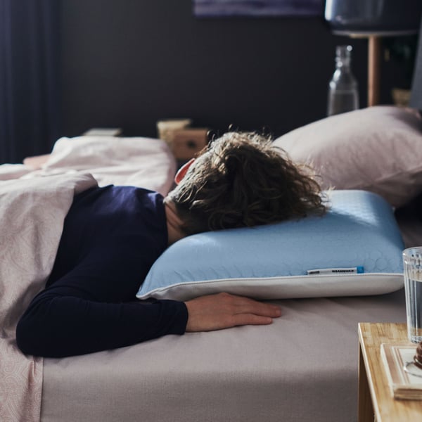 Dans une chambre sombre, une personne en pyjama bleu dort sur le ventre, la tête posée sur un oreiller ergonomique KVARNVEN.