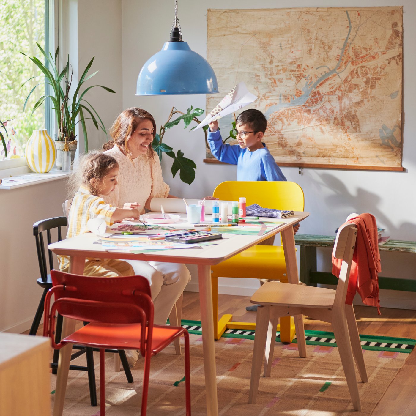A woman and child paint at a LISABO table with colourful chairs, while another child nearby holds a paper airplane.