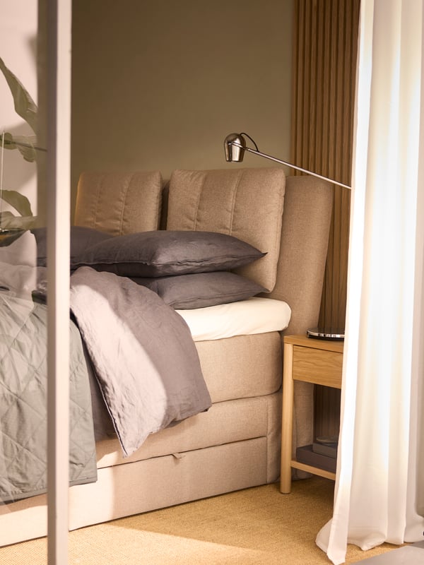 The top end of a beige LILLFJÄLLET double bed with storage covered with dark grey DYTÅG bed linen in a sunny room.