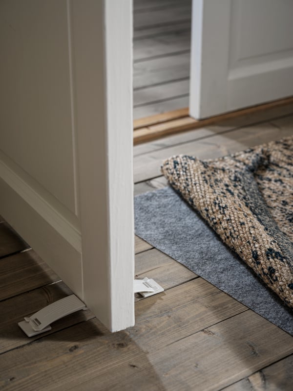 An open door held in place by a white PATRULL door stop. By the door is rug, a lifted corner revealing anti-slip underlay.