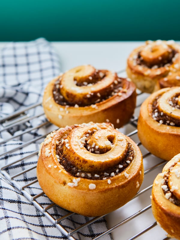Several KAFFEREP cinnamon buns on a metal oven grill, on a white surface above a tea towel with checked design.