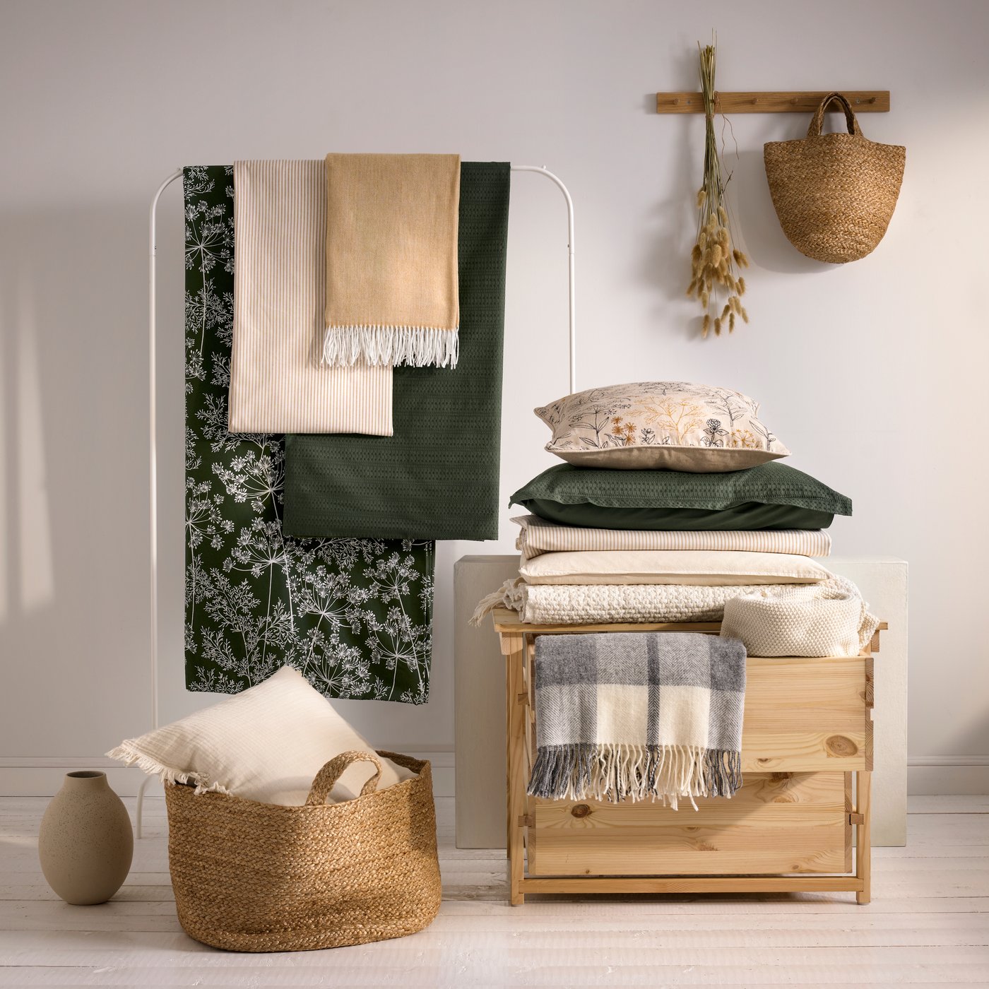 A studio scene with blankets on a clothes rack, a basket and a wooden chest of drawers pilled with quilts and pillows.