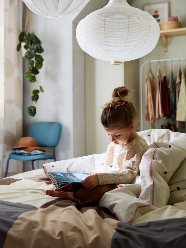 Fillette enveloppée d’un plaid lisant sur un lit habillé d’une housse de couette VÄNKRETS à pois blancs et roses et surmonté de deux suspensions blanches.