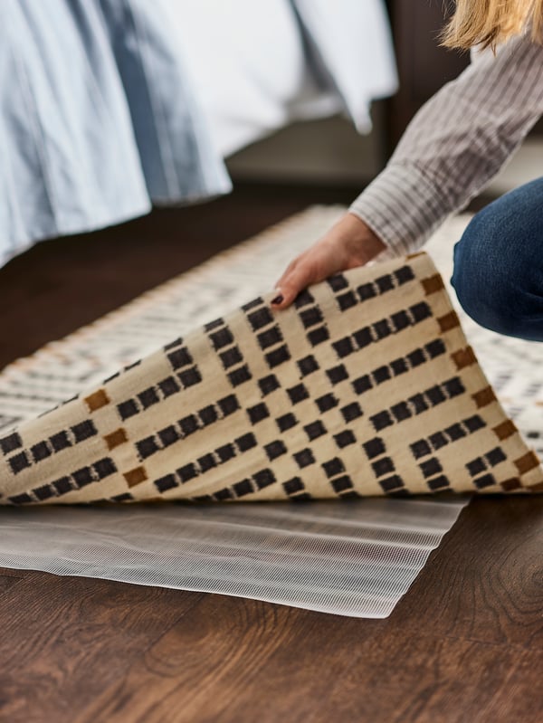 Une main soulève le coin d’un tapis VAMDRUP multicolore tissé à plat près d’un lit, montrant une thibaude antidérapante en dessous.