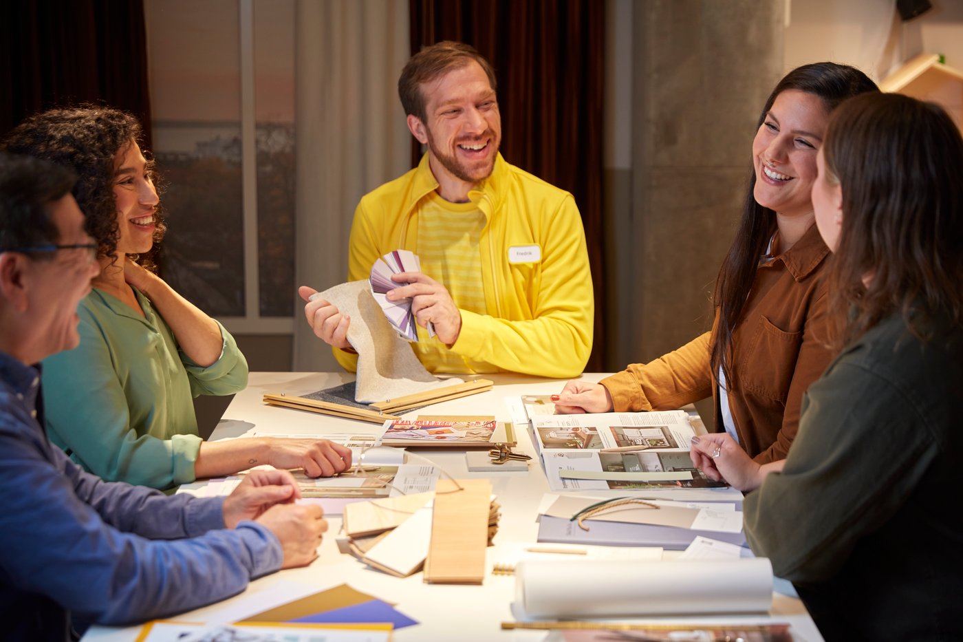 En smilende IKEA medarbejder viser tekstil- og farveprøver til deltagerne i en workshop.